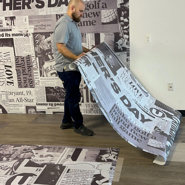 Man applying large custom vinyl prints with adhesive backing featuring newspaper-style graphics on wall and floor