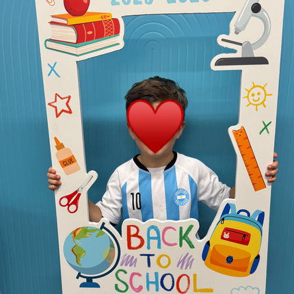 Child holding a back to school photo frame backdrop with school supplies and 2025-2026 text