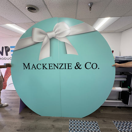 ROUND Cut to Shape Foam Backdrop featuring a decorative bow and Mackenzie & Co branding in teal